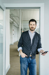 Porträt eines Geschäftsmannes, der ein Telefon in der Hand hält, während er an der Tür eines Büros steht - CAVF25209