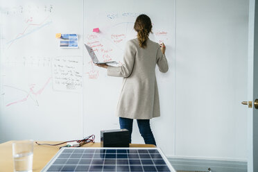 Rear view of businesswoman writing on whiteboard while working over solar panels in office - CAVF25214