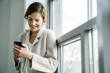 Geschäftsfrau, die ein Smartphone benutzt, während sie im Büro am Fenster sitzt - CAVF25215