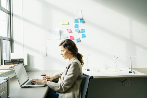 Seitenansicht einer Geschäftsfrau, die einen Laptop benutzt, während sie an einem Windradmodell auf dem Schreibtisch arbeitet - CAVF25221