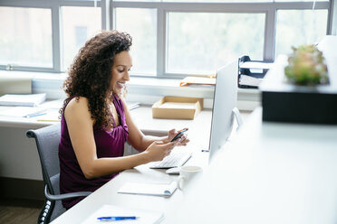 Lächelnde Geschäftsfrau, die ein Mobiltelefon benutzt, während sie am Schreibtisch im Büro sitzt - CAVF25236