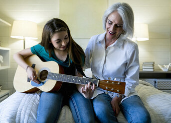 Grandmother looking at granddaughter playing guitar while sitting on bed - CAVF25317