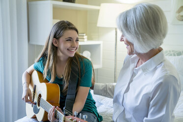 Happy granddaughter playing guitar while sitting by grandmother in bedroom - CAVF25320