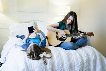 Girl using smart phone while sister playing guitar on bed at home - CAVF25329