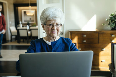 Senior woman using laptop computer at home with senior man in background - CAVF25373