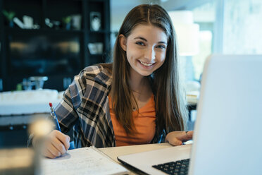 Portrait of happy teenage girl doing homework using laptop computer at home - CAVF25376