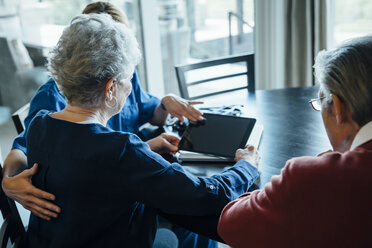 Hauspflegerin diskutiert mit einem älteren Ehepaar am Esstisch über einen Tablet-Computer - CAVF25389