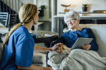 Ältere Frau, die im Wohnzimmer einen Tablet-Computer hält und die häusliche Pflegekraft anschaut - CAVF25400