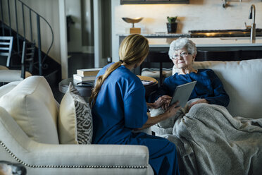 Home caregiver discussing over tablet computer with senior woman in living room - CAVF25401