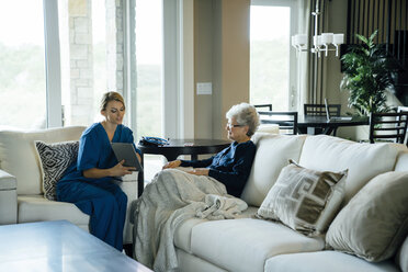 Home caregiver showing tablet computer to senior woman while sitting on sofa in living room - CAVF25406