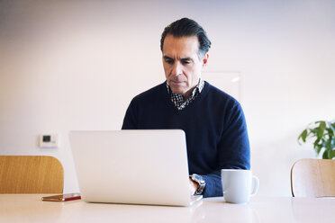 Ernster Mann schaut auf einen Laptop-Computer, während er an einem Tisch an der Wand sitzt - CAVF25417