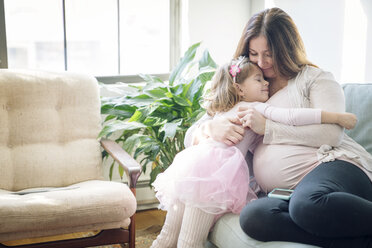 Glückliche schwangere Frau, die ihre Tochter umarmt, während sie zu Hause auf dem Sofa sitzt - CAVF25427