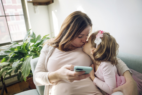 Mutter küsst Tochter und hält Smartphone zu Hause, lizenzfreies Stockfoto