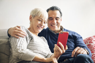 Lächelndes älteres Paar schaut auf sein Smartphone, während es zu Hause auf dem Sofa sitzt - CAVF25433