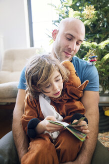 Vater sitzt mit seinem kostümierten Sohn zu Hause vor dem Weihnachtsbaum - CAVF25437