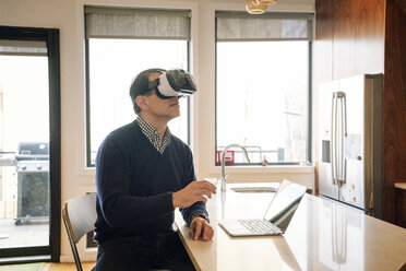 Man wearing virtual reality simulator sitting at table in kitchen - CAVF25448
