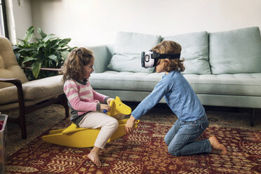 Junge, der einen Virtual-Reality-Simulator trägt, spielt mit seiner Schwester, die zu Hause auf einem Schaukelpferd sitzt - CAVF25454