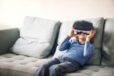 Happy boy wearing virtual reality simulator sitting on sofa at home - CAVF25455