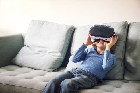 Glücklicher Junge mit Virtual-Reality-Simulator auf dem Sofa zu Hause - CAVF25455