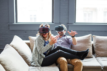 Happy coworkers wearing virtual reality simulators while sitting on sofa in office - CAVF25582