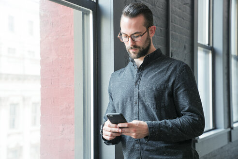 Geschäftsmann, der sein Smartphone benutzt und sich im Büro an die Wand lehnt - CAVF25599