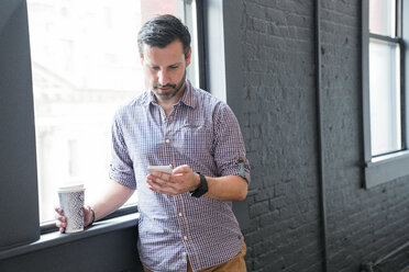 Businessman using smart phone while standing by window in office - CAVF25601