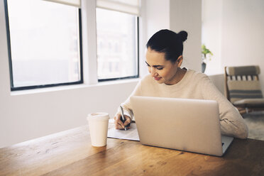 Businesswoman writing while sitting with laptop computer at desk in office - CAVF25611