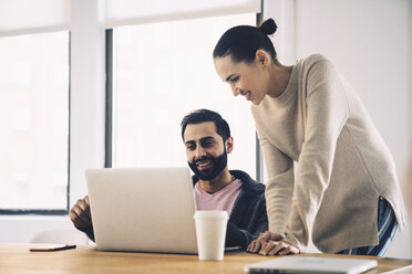 Lächelnde Kollegen, die beim Diskutieren im Büro auf einen Laptop schauen - CAVF25620