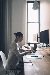 Side view of businesswoman using laptop computer while sitting at desk in office - CAVF25623