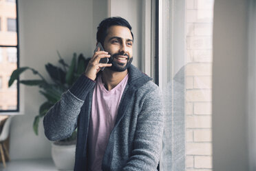 Lächelnder Geschäftsmann, der mit seinem Smartphone spricht, während er auf der Fensterbank im Büro sitzt - CAVF25629