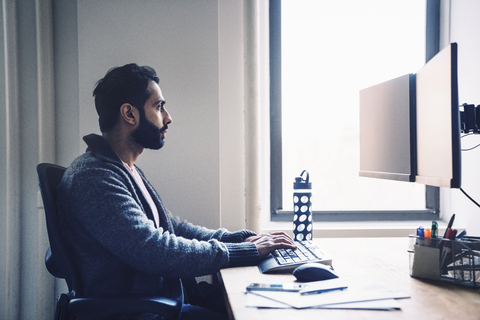 Seitenansicht eines Geschäftsmannes, der einen Desktop-Computer benutzt, während er am Schreibtisch im Büro sitzt, lizenzfreies Stockfoto
