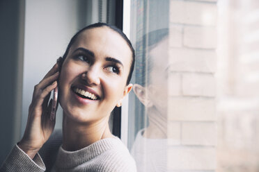 Happy woman looking through window while talking on mobile phone at home - CAVF25636