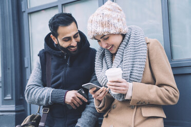 Happy couple using mobile phone while leaning by building in city - CAVF25642