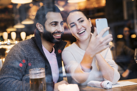Happy woman with boyfriend using mobile phone seen through restaurant window stock photo