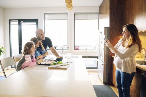 Schwangere Frau fotografiert Familie am Küchentisch zu Hause - CAVF25653