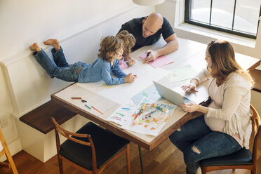 Blick von oben auf den Vater, der seinen Kindern beim Zeichnen hilft, während die Frau am Tisch einen Laptop benutzt - CAVF25657