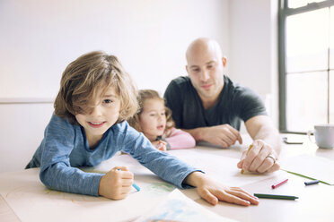 Portrait of boy drawing while father assisting daughter at table in home - CAVF25659