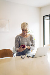 Happy senior woman using mobile phone while sitting at kitchen table in home - CAVF25664