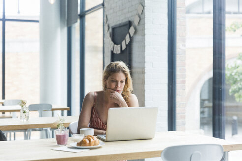 Frau mit Hand am Kinn, die einen Laptop benutzt, während sie in einem Café sitzt - CAVF25868