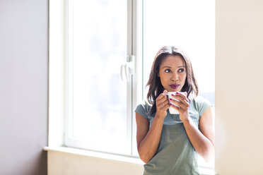 Nachdenkliche Frau trinkt Kaffee, während sie zu Hause am Fenster steht - CAVF25875