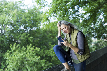 Woman with hand in hair using smart phone while listening music by retaining wall against branches - CAVF25919