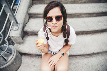 Hochformatiges Porträt einer Frau mit Sonnenbrille, die auf einer Treppe Fruchtsaft trinkt - CAVF25944