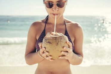 Frau mit Sonnenbrille und Bikini beim Trinken von Kokosnusswasser am Strand gegen das Meer - CAVF25945