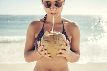 Woman wearing sunglasses and bikini while drinking coconut water at beach against sea - CAVF25945