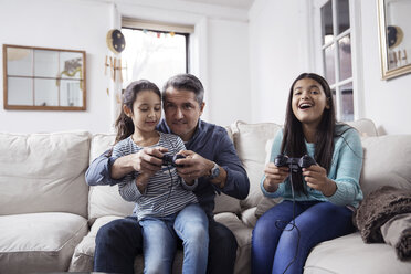 Girls with father playing video game while sitting on sofa at home - CAVF26059