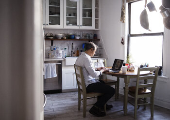 Seitenansicht eines Mannes, der einen Laptop benutzt, während er am Esstisch in der Küche sitzt - CAVF26073