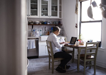 Side view of man using laptop computer while sitting at dining table in kitchen - CAVF26073
