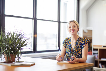 Porträt einer lächelnden Geschäftsfrau mit Mobiltelefon, die am Tisch im Büro sitzt - CAVF26114