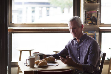 Mature man using phone while having croissants and coffee at table in cafe - CAVF26155