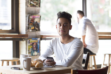 Portrait of young man holding mobile phone having croissant and coffee at cafe - CAVF26179